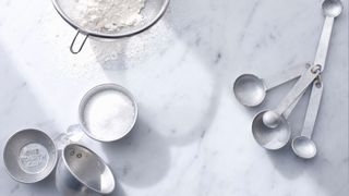 Metal measuring spoons on a marble worktop, beside a sieve full of flour and a cup of sugar