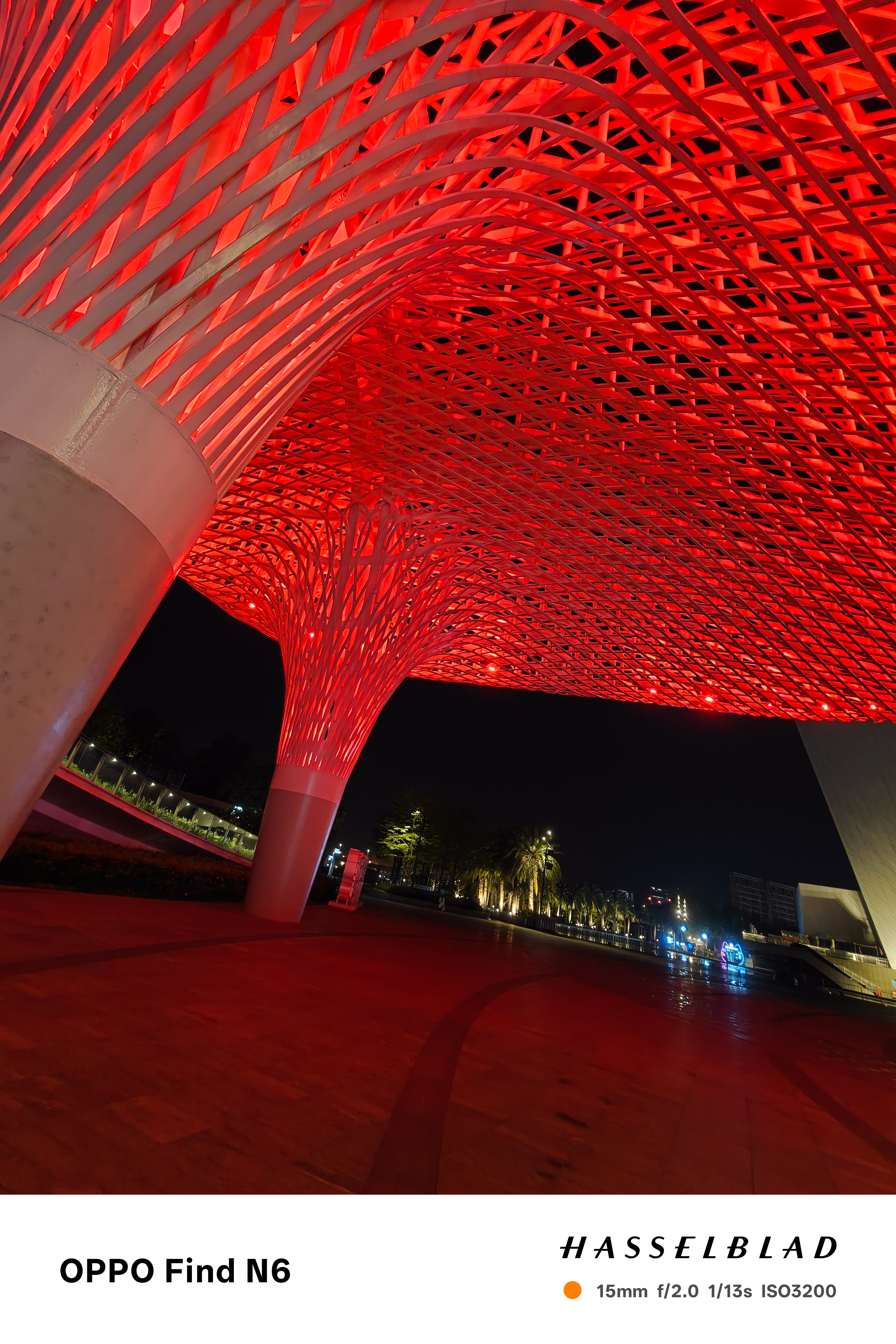 An ultra-wide-angle night shot from beneath a massive, tree-like architectural structure made of a complex red lattice. The structure is brilliantly illuminated from within, casting a red glow over the dark, reflective ground of the outdoor plaza.