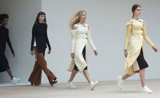 Four models on catwalk, one in a black dress, one in a black top and brown trousers, and two in cream coats