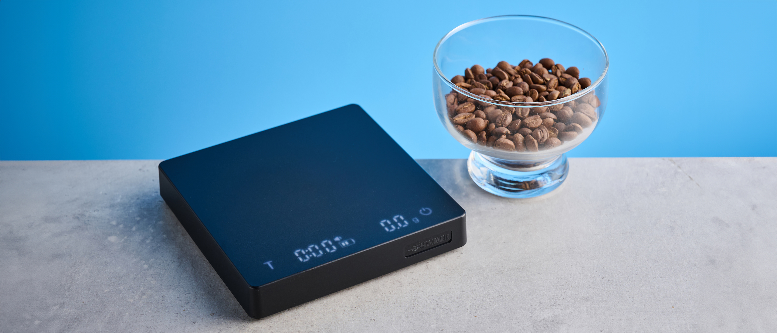 the mhw-3bomber cube coffee scale 2.0 in black photographed against a blue background showing its button controls and dial surface