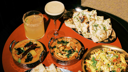 An orange table at Tamila, London with steel dinnerware serving naan, two curries, a vegetable side dish, and an espresso martini, and spicy cocktail in glassware