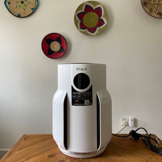 Shark NeverChange 5 Air Purifier standing on a wooden table in front of a white wall with colourful wicker baskets on it