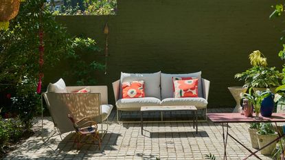 Image of an outdoor patio with a green wall at the back of the space. There is a small, light gray outdoor sofa and a matching accent seat, and a few small tables and plants around the patio.