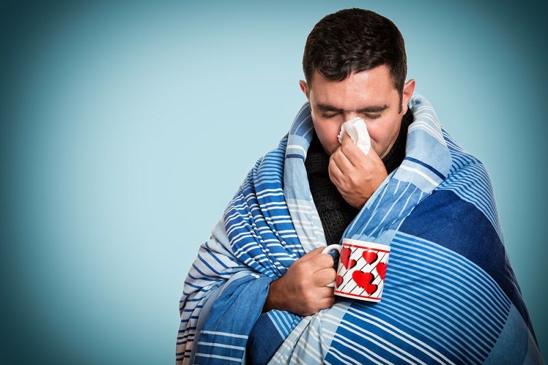 An ill man blowing his nose and holding a cup of tea.