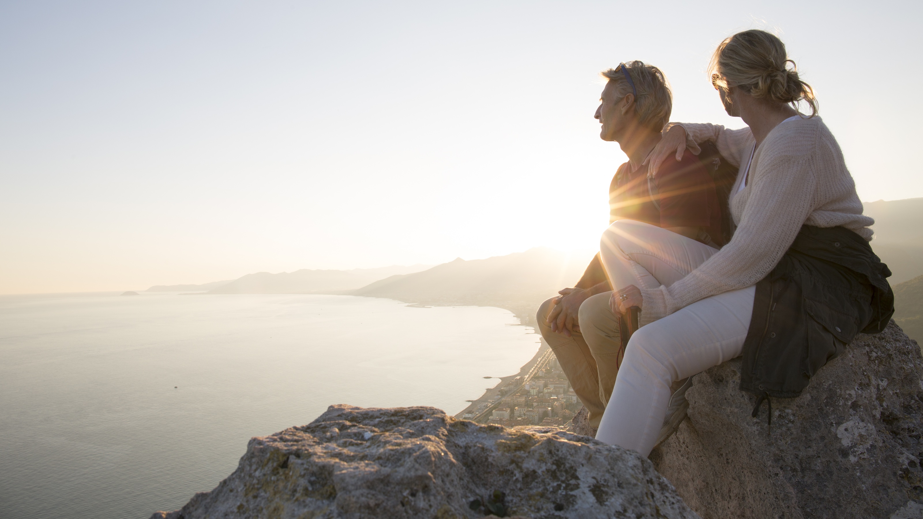 A couple take time to enjoy the sunset over the water while on a mountain hike.