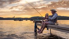 An older man smiles as he fishes on a dock with his grandson at sunset.