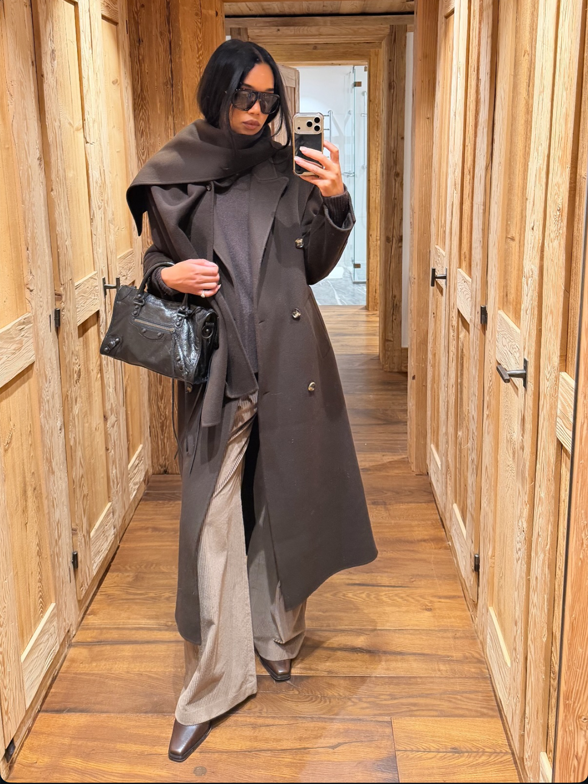 Laura Harrier wearing a brown coat, beige corduroy pants, and brown boots with a black Balenciaga City bag.