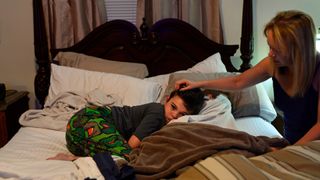 A child wearing a gray shirt and patterned green pants curls up on a bed with his mother, a blond woman with long straight hair, caressing his head