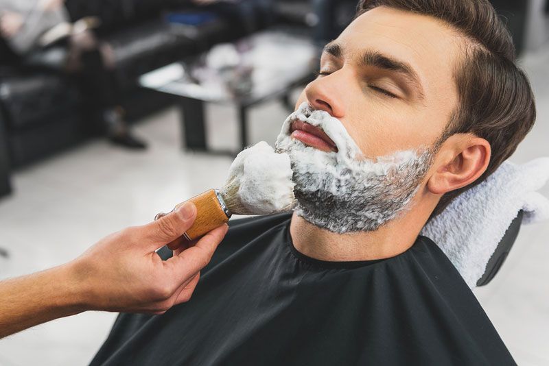 A barber applying shaving cream with a shaving brush to a man&#039;s face.