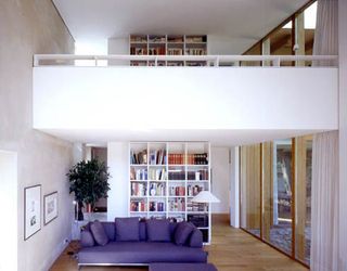 Interior room, wooden floor, white walls, white viewing gallery wall, white central book case with books and other items, purple soft sofa, picture frames, right side glass framed wall, potted tree in the left corner, floor standing lamp with white shade, white curtains