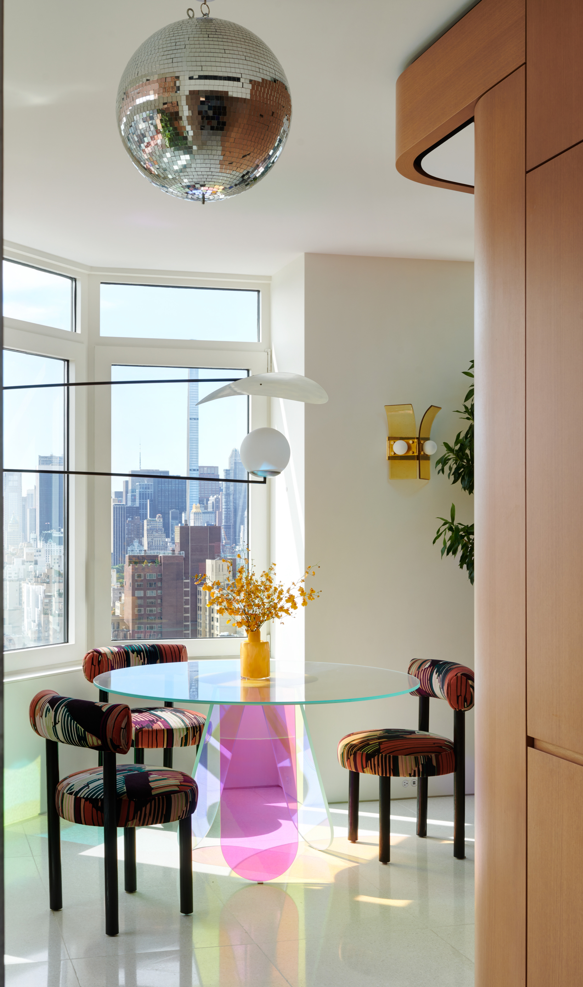 Image of a white, modern dining nook with a glass table and dark patterned chairs. There are lots of windows on the walls and a disco ball hanging overhead.