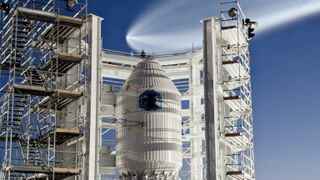 A white conical vessel with round window near the top stands surrounded by white scaffolding under a dark blue evning sky. Above, a giant, jelly-fish like plume trails a rocket launching through the sky.