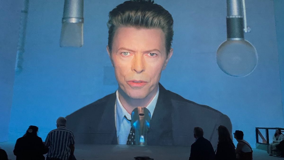 A large projection of David Bowie in a suit and tie, with people sitting in front of the image at Lightroom, Kings Cross, as part of the David Bowie: You're Not Alone exhibition.