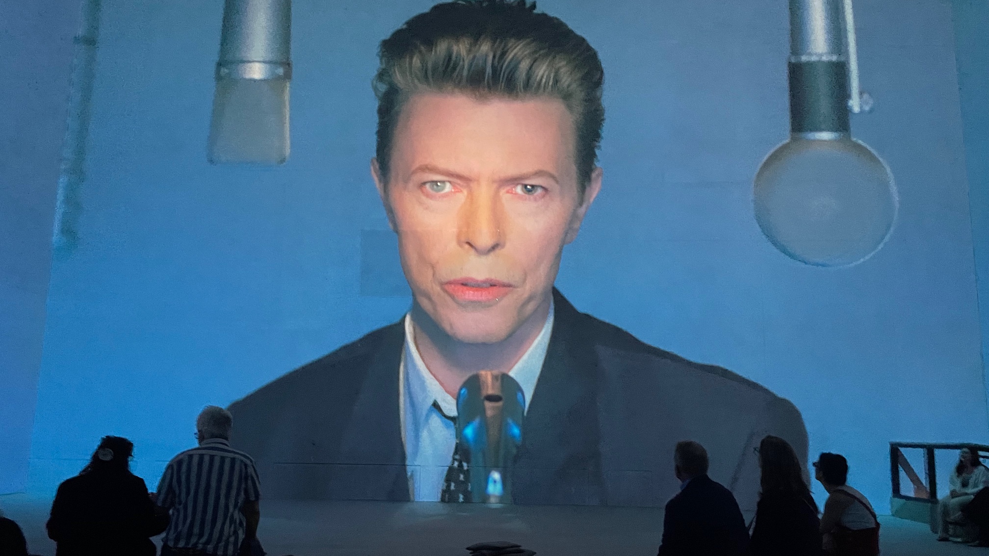A large projection of David Bowie in a suit and tie, with people sitting in front of the image at Lightroom, Kings Cross, as part of the David Bowie: You're Not Alone exhibition.