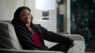 Simone Biles smiling while sitting on the couch.