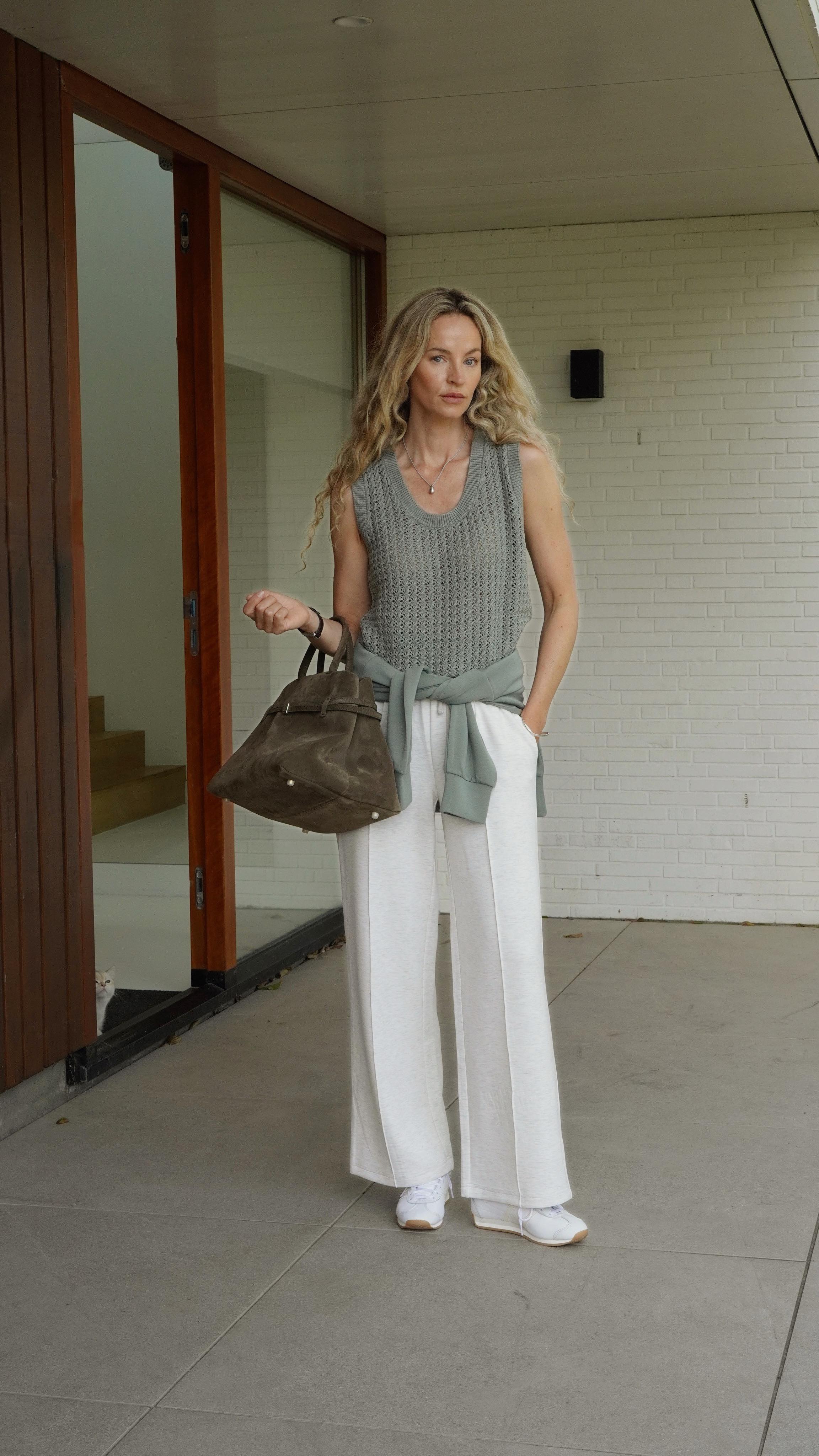 An image of @anoukyve wearing a grey scoop neck sweater vest with white wide-leg pleated trousers, a suede Manu Atelier bag and white trainers.