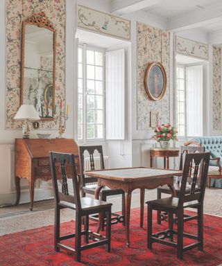 French dining table and chairs with living space to right, antique furniture, rug, large gold framed mirror and portraits, writing desk, window shutters, tiled floor, floral wallpaper with birds