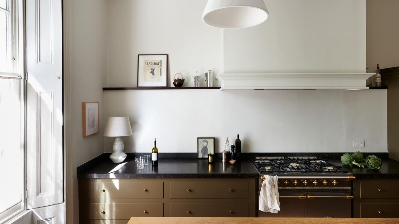 A Georgian terrace in Regent&#039;s Park with olive painted drawers and a dark countertop. A gas cooking stove is seen on the right, as well as open shelving on the left side with artwork on the wall 