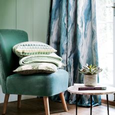 Green velvet cocktail chair in front of a green painted panelled wall with a stack of green cushions and throws on the chair