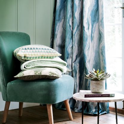 Green velvet cocktail chair in front of a green painted panelled wall with a stack of green cushions and throws on the chair