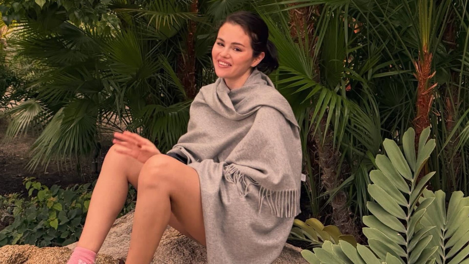 Selena Gomez sits on a large rock surrounded by trees and shrubbery wearing a scarf knit with Uggs and pink socks.