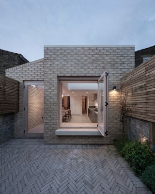 A backyard to an apartment with grey brick flooring, shrubbery, wooden walls, a glass door and a large open window.