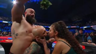 Ricochet and Samantha Irvin kissing under a mistletoe held by Braun Strowman on SmackDown