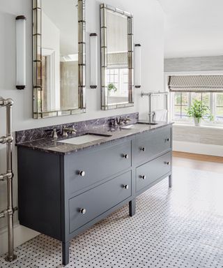 Large bathroom with dark gray sink and storage unit, black marble countertop, twin sinks with matching metal mirrors hanging above, slender wall lights hanging either side, tiled flooring