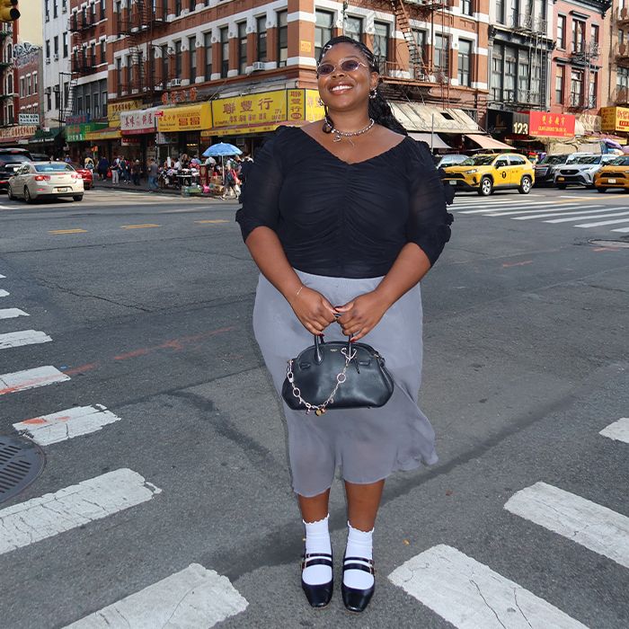 chichi wears black blouse and gray sheer skirt with black top handle coach bag and white socks with black shoes while posing in the streets of NYC