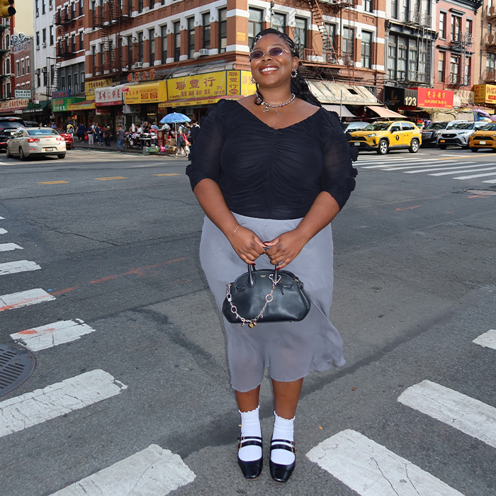 chichi wears black blouse and gray sheer skirt with black top handle coach bag and white socks with black shoes while posing in the streets of NYC
