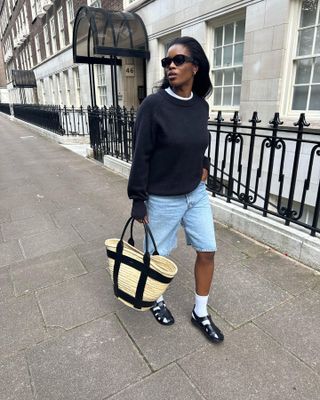 Woman on street wears black sweatshirt, blue shorts, black sandals, carries basket bag