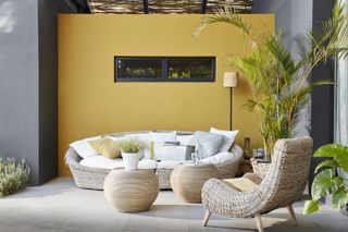 Curved wicker outdoor patio furniture in a yellow and gray painted scheme with large potted plants.