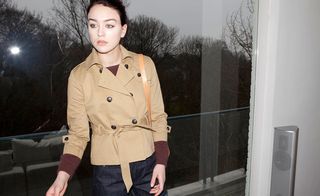 Dusk image of a Female model stood on a low glass framed balcony, wearing black a beige jacket with deep red jumper, jeans, grey sky, surrounding trees and grass area in the background