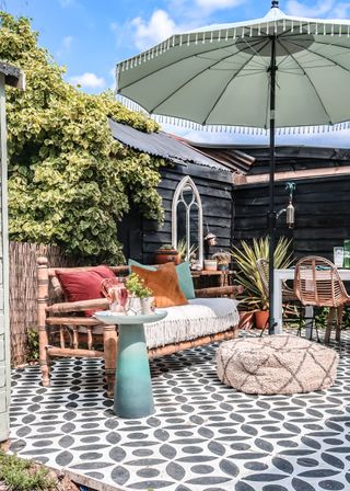 A black and white patterned patio space with Morrocan-style furniture including a tasselled umbrella, pouffe and pink, orange and blue soft furnishings.