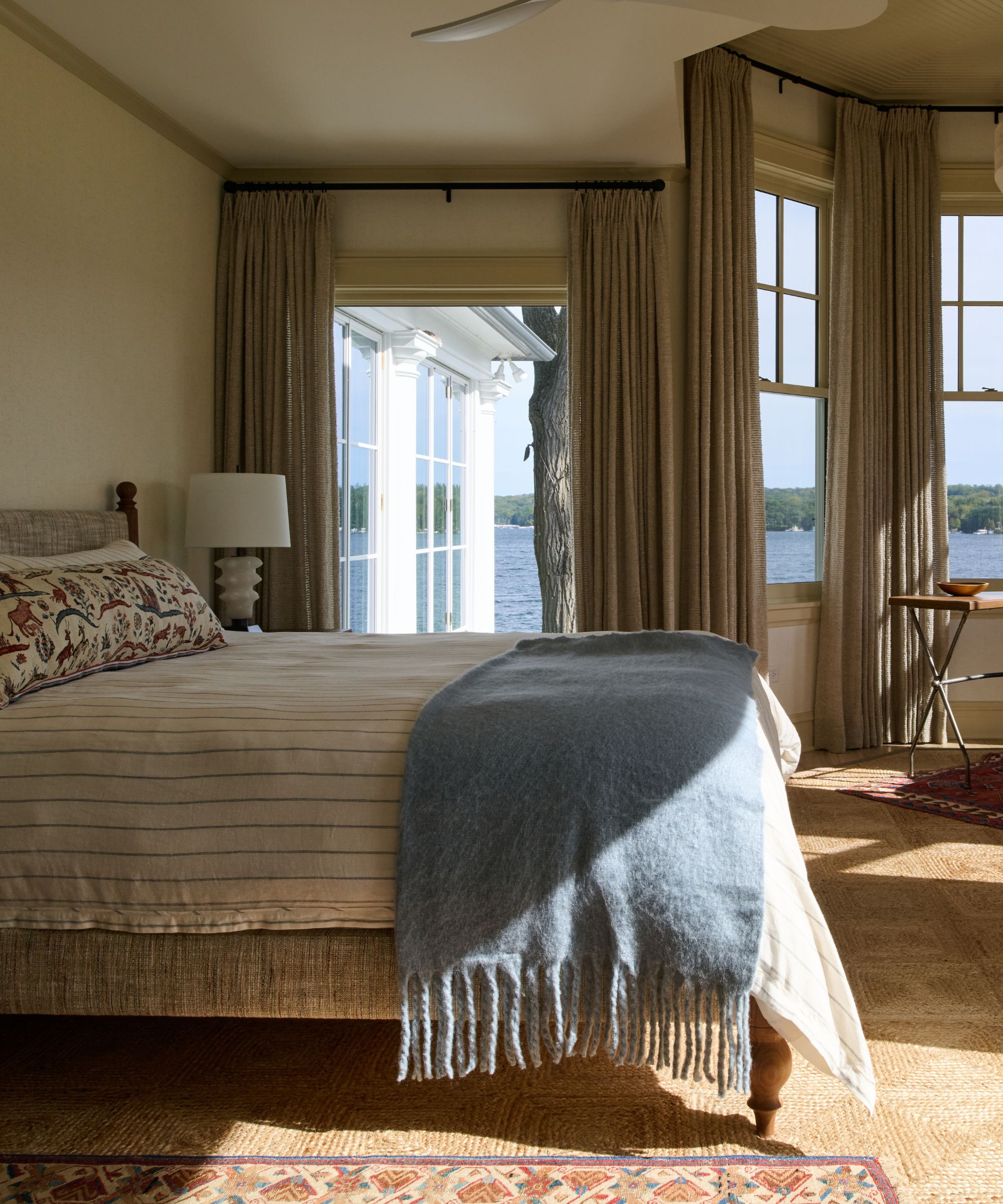 A bedroom with a neutral color scheme and large windows with a powder blue throw on the bed.