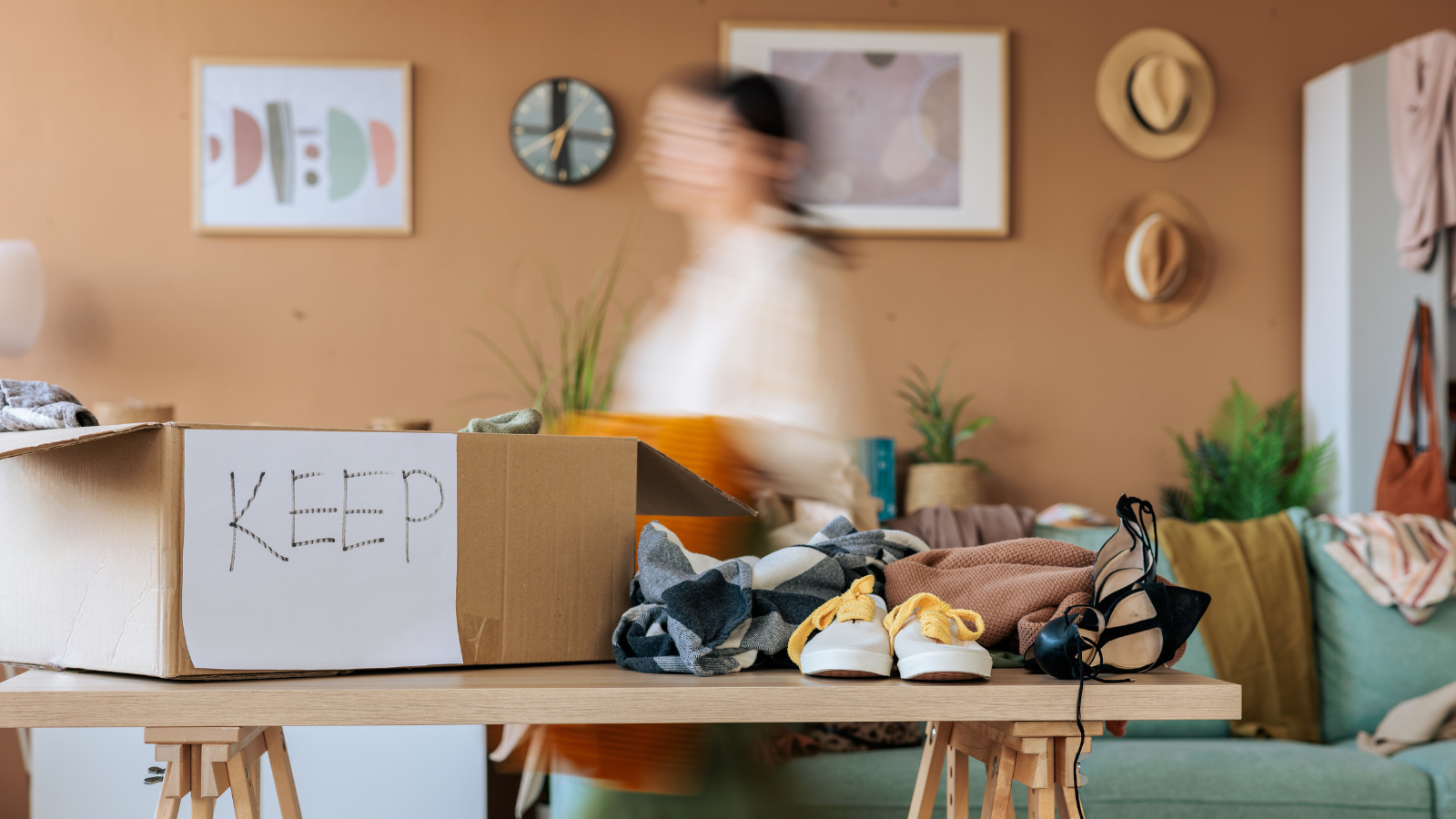 A cardboard box with a keep sign on a table next to clothes and shoes and a person walking in the background