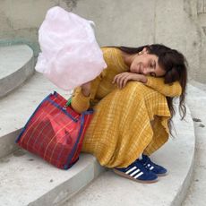 Influencer @dianekari sits down on stairs outside wears a yellow dress with navy adidas trainers. She holds a stick of candy floss and rests her head on her arm.