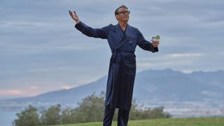 Jason Isaacs in a blue dressing gown as Cary Grant stands in front of a background of mountains and sea holding a drink with his arms out in Archie.