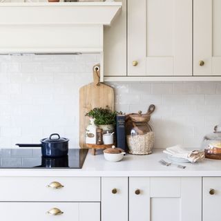 Kitchen with cream cupboards, a white worktop, and white tiles behind the hob. The worktop is covered in cooking accessories