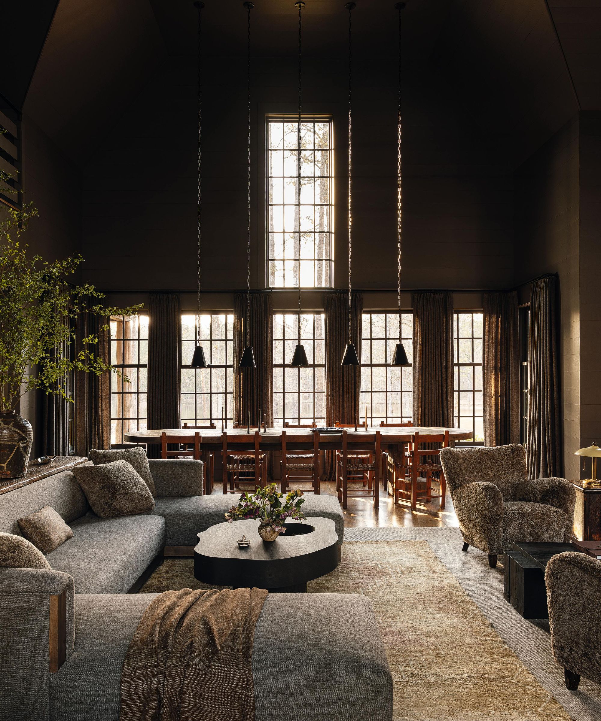 a moody farmhouse with wood clad walls, large windows, a dining table, a large grey section couch and shearling arm chairs