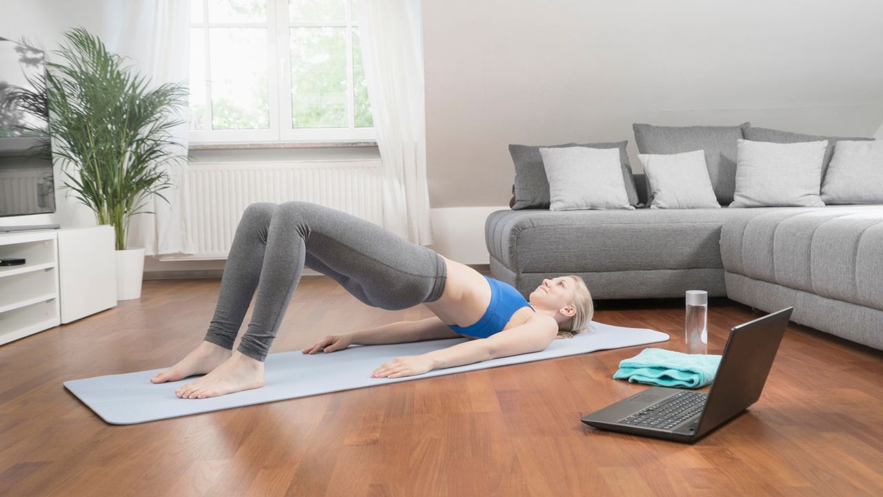 woman performing a pilates bridge on an exercise mat in a living room setting with a laptop to the side of her and a grey corner sofa behind her.