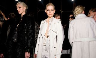 Three female models wearing looks from Roberto Cavalli's collection. One model is wearing a black fur jacket. Next to her is a model wearing a white blouse, trousers, coat and long necklace. And the third model is wearing a off-white coloured belted coat. There are other people in the background