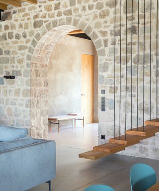 living area with exposed stone wall, gray sofa, wooden floating stairs and aqua chairs i