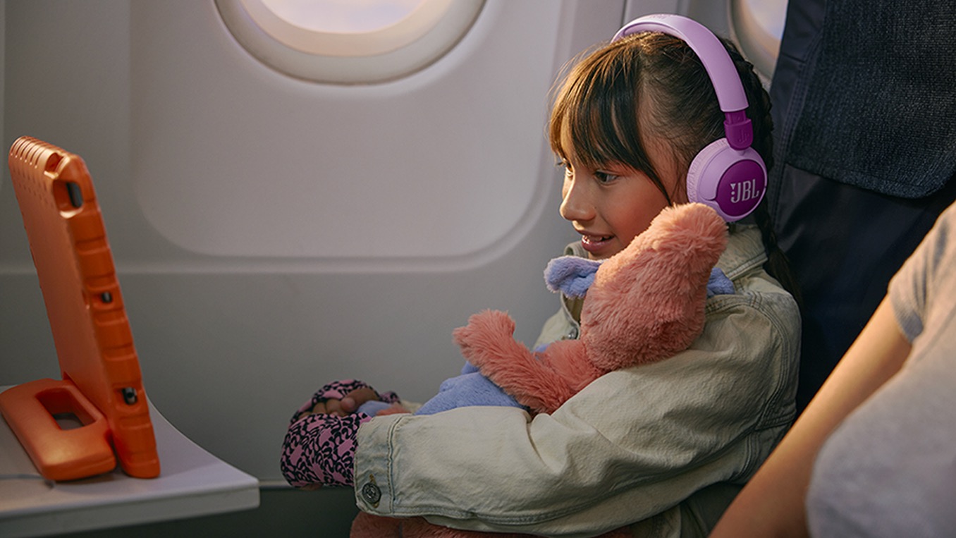A girl hugging a soft toy in a plane seat while wearing a pink pair of JBL Junior 320BT headphones and watching a tablet.