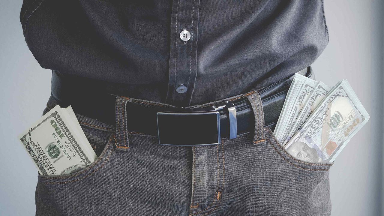 A man has stacks of $100 bills sticking out of both pants pockets.