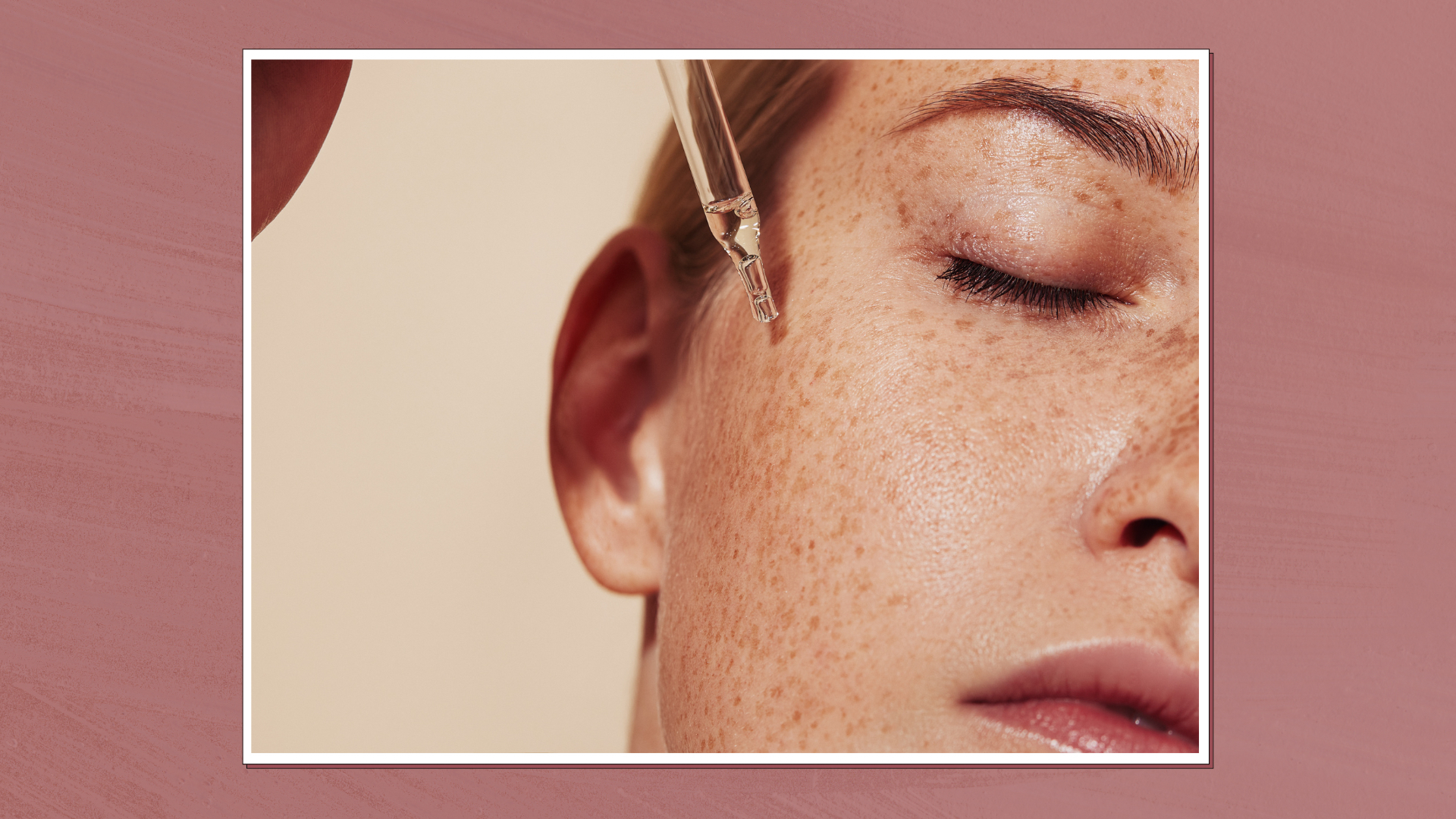 Close up image of a woman's face while she drips pipette serum onto her skin, in a white frame set against a dark pink background 
