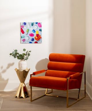 An orange velvet chair in a corner with a gold side table, small plant, and a patterned print on the wall