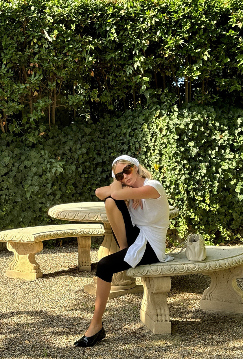 How to pack for Italy is shown in a picture of a woman wearing a white headscarf, black sunglasses, a white t-shirt, black capri pants, and black ballet flats