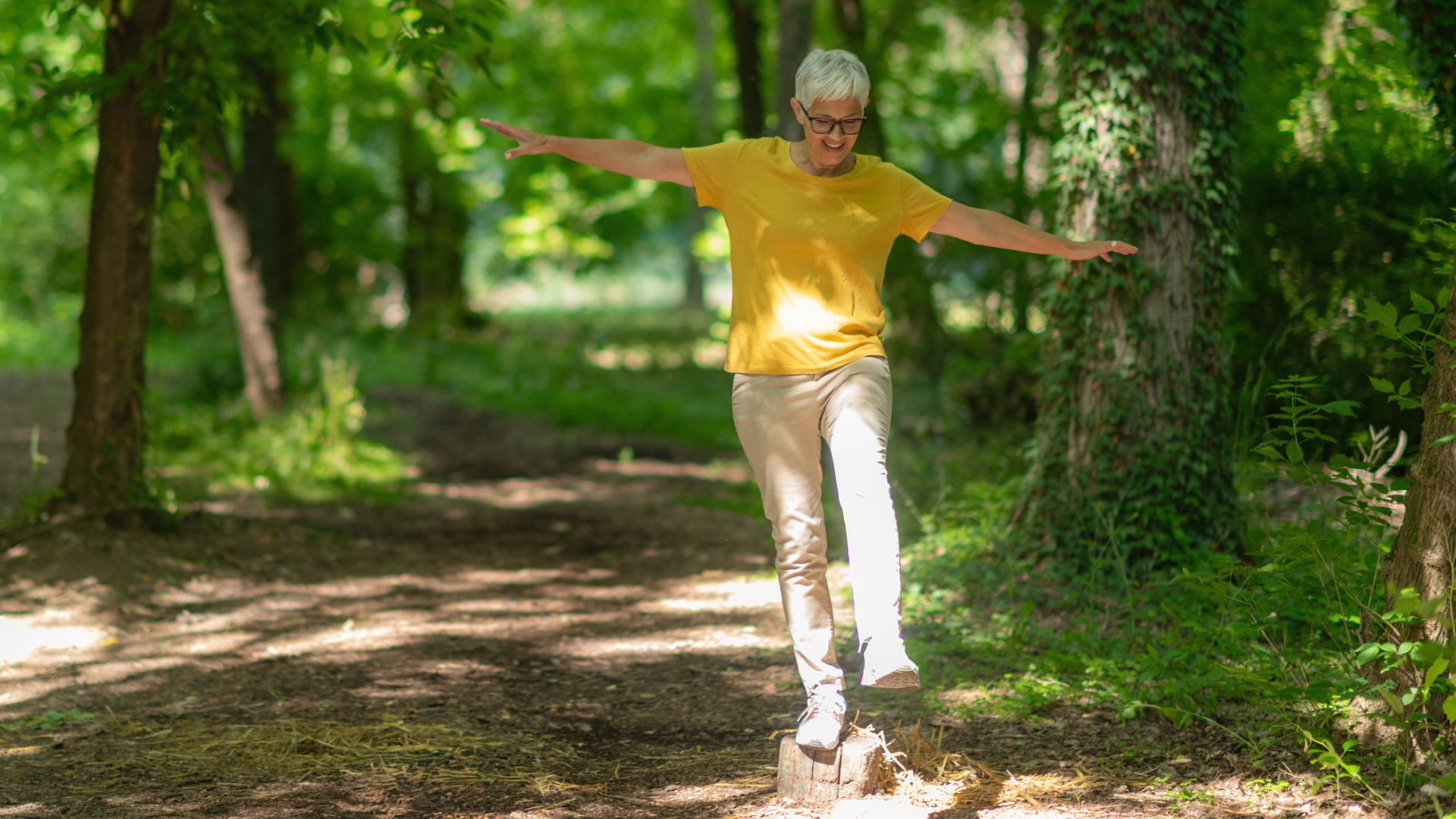woman outside in a forest setting balancing on one leg on a small tree stump with arms out to her sides. she's wearing a yellow tshirt and cream trousers. she has white short hair and wearing glasses. 