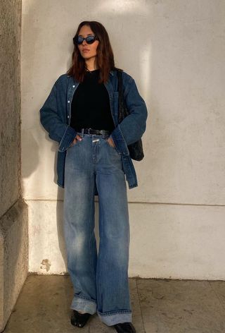 A winter travel outfit is shown in a photo of a woman standing outside wearing a denim shirt over a black crewneck sweater styled with a black belt, relaxed jeans, black ballet flats, a black hobo bag, and sunglasses